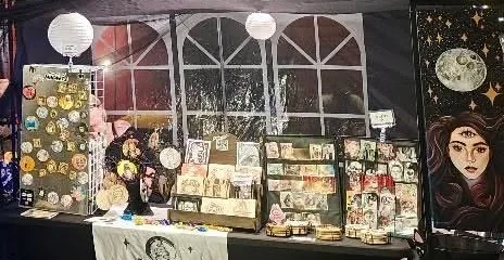 Sara's market table, with hand painted magnets on a pegboard, racks of prints and cards, and a large painted moon portrait