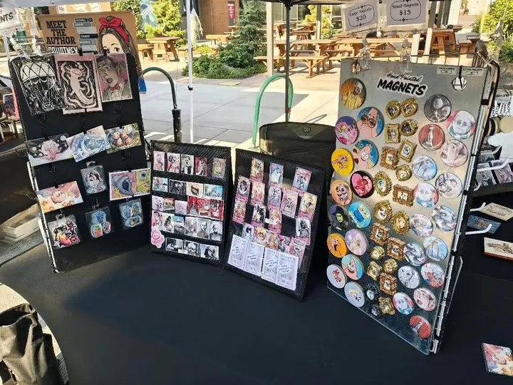 Racks of prints and hand painted button magnets on a board headed Magnets, on her table in daylight