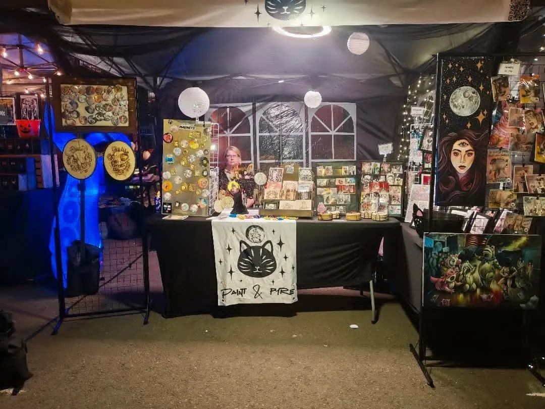 The stall after dark under a dark canopy, lit from inside, with framed originals on the side walls and a painted moon portrait on the right