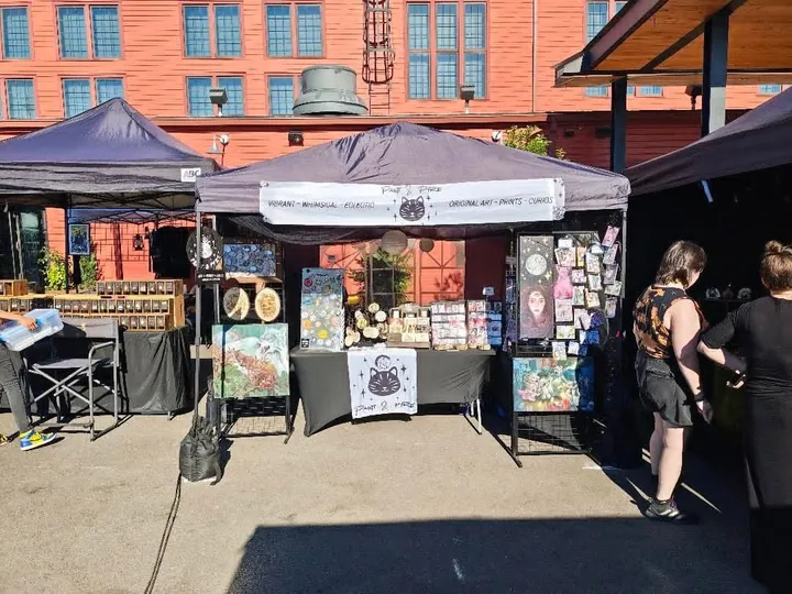 The black canopy set up on a street in front of a red brick building, with two customers looking at the prints