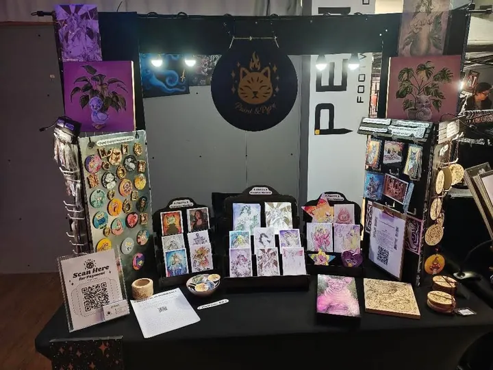 Her table indoors, lit purple, with a spinner of button magnets, racks of prints and a hanging banner with her cat mark