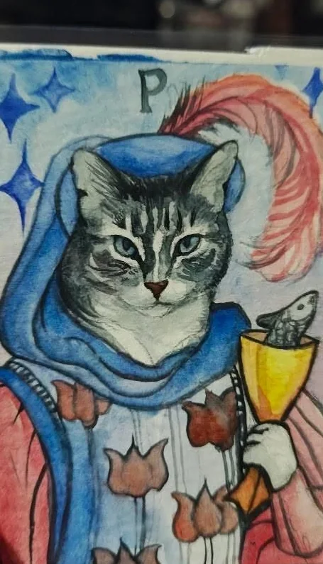 A hand painted tarot card lettered P, a grey tabby cat in a blue hood and a plumed hat, holding a gold cup with a fish in it