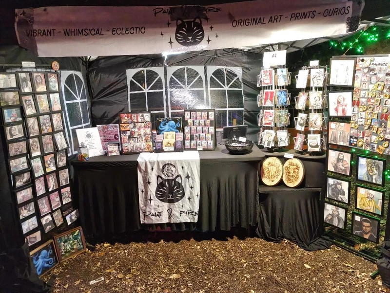 Inside the Paint & Pyre booth, with a black cat banner, portrait displays and a table of small artwork