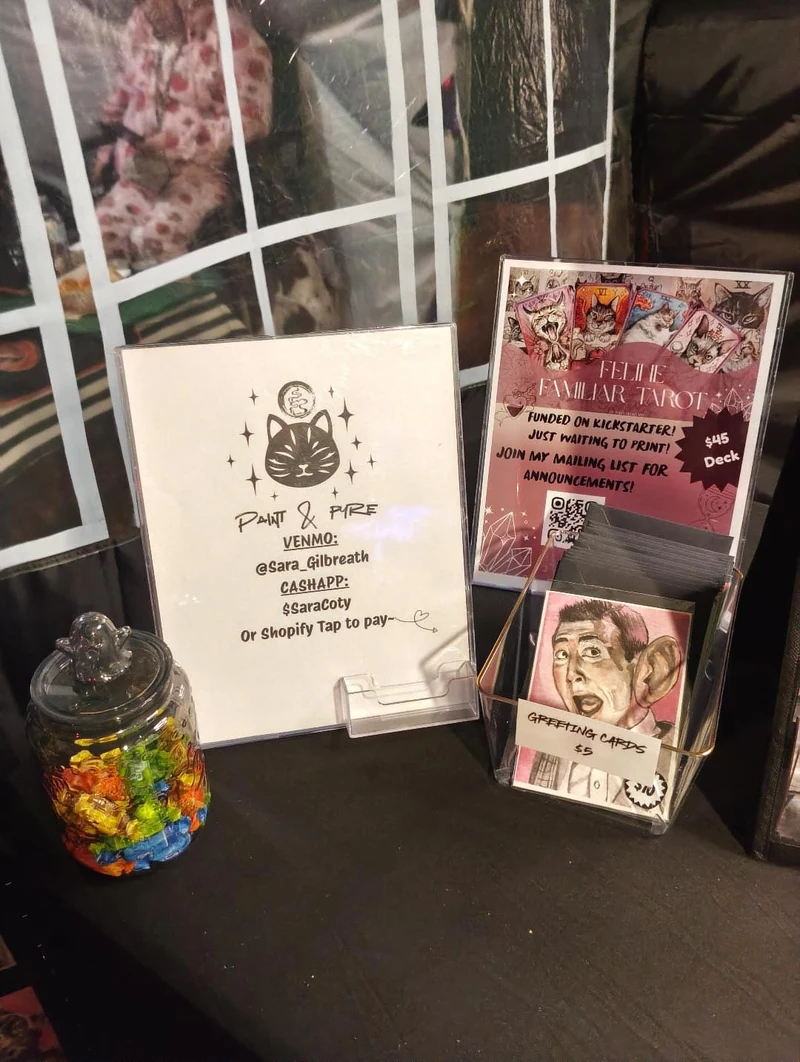 Paint & Pyre’s cat logo and information cards beside the card display on a black market table