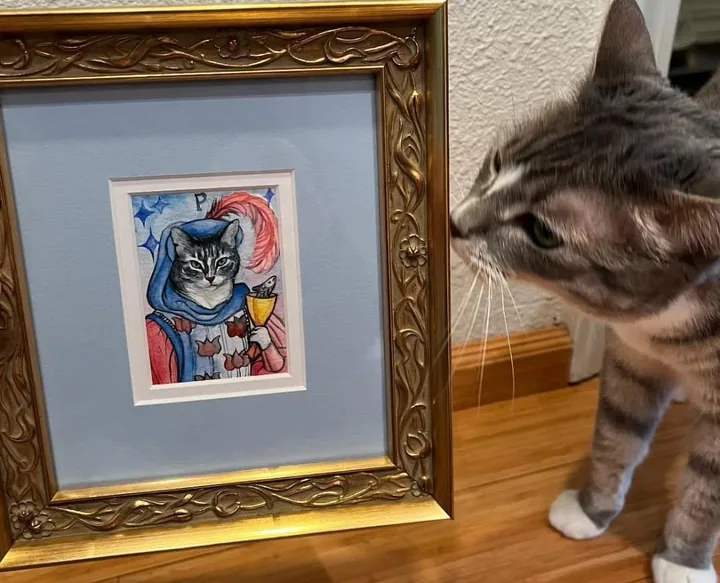 A tabby cat sniffing her own painted portrait, framed in gold and standing on a wooden floor
