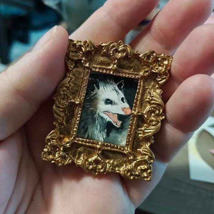A painting of an opossum about the size of a postage stamp, in an ornate gold frame, held in the palm of a hand