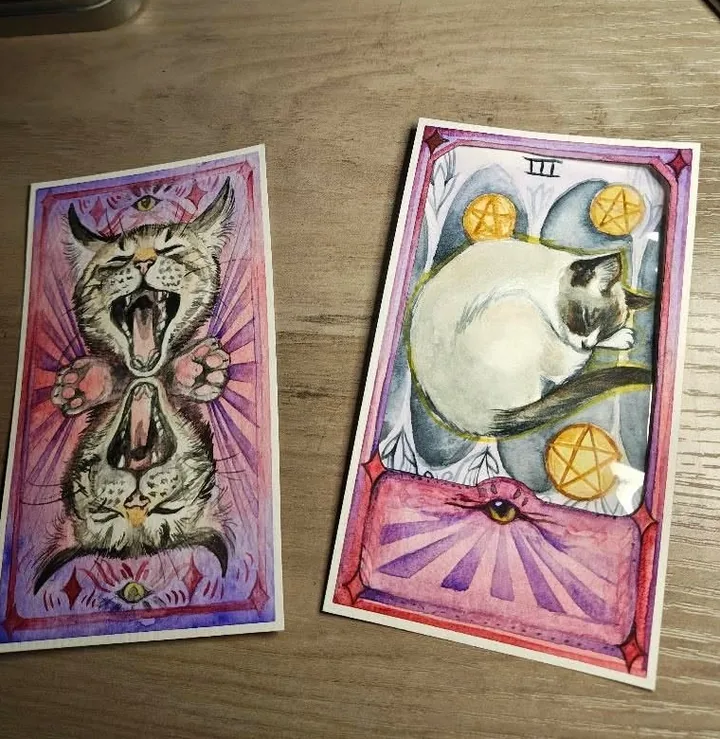 Two printed tarot cards lying on a wooden desk, one a yowling cat, one a siamese cat asleep among five pentacles