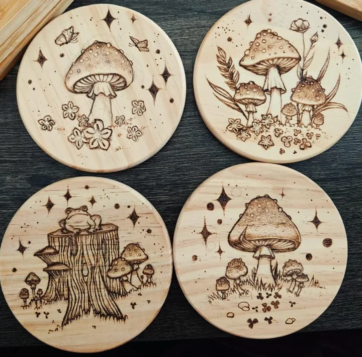 Four wood burned coasters laid out on a dark table, three with mushrooms and one with a frog on a tree stump